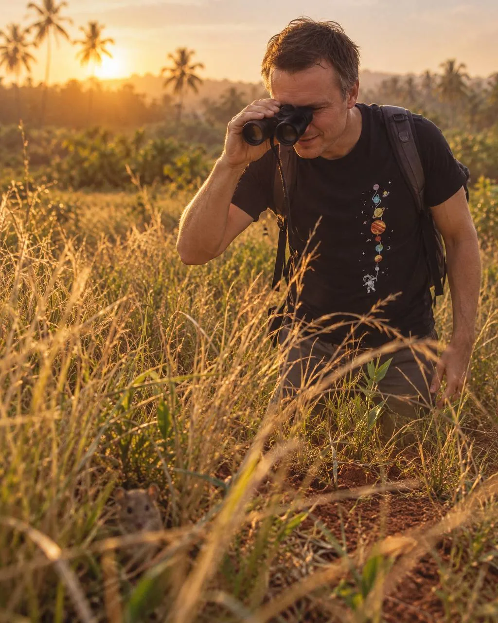Dmitry in tall grass at sunset, looking through binoculars, with a vole sitting at his feet